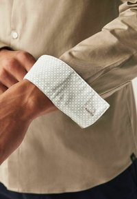 Cămașă de culoare maro deschis cu manșete albe cu model. Manșeta are un design în carouri și detalii din hardware argintiu. Textură de țesătură netedă.