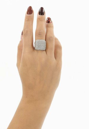 Dłoń z brązowym i panterkowym lakierem do paznokci, nosząca duży, kwadratowy srebrny pierścionek wysadzany małymi kryształkami na palcu środkowym.