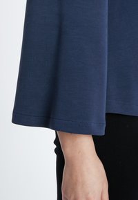 Marineblaue Langarmbluse mit glatter Textur und weiten, ausgestellten Ärmeln. Der Saum ist mit feinen Nähdetails versehen; getragen mit schwarzen Unterteilen.