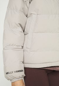Svetle šedá péřová bunda s prošívanými sekcemi, elastickými manžetami a logem "Helly Hansen" na rukávu. Měkká textura a oversize střih.