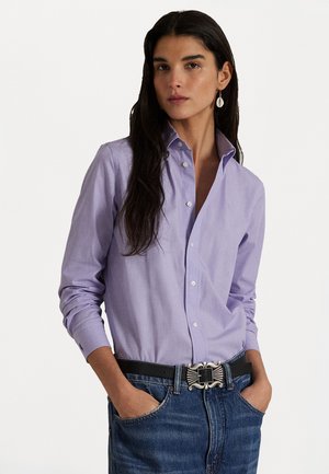 Camicia a maniche lunghe di un leggero colore viola con una texture morbida e un colletto strutturato, abbinata a jeans di denim scuri e una cintura decorativa in argento.