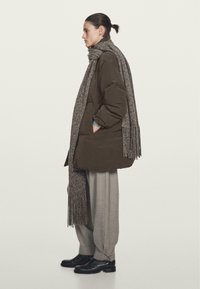 Pufferjakke i mørk olivengrøn, med et struktureret gråt tørklæde, løse beige bukser og sorte ankelstøvler, stående mod en lys baggrund.