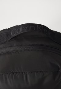 Černá péřová taška s uzávěrem na zip, texturovanou látkou a vyšitým logem Calvin Klein na plochém popruhu. Minimalistický design.