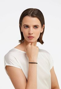 Fekete fém karkötő domború felirattal, kör alakú forma, sima textúra, kézen viselve, fehér felsővel párosítva.