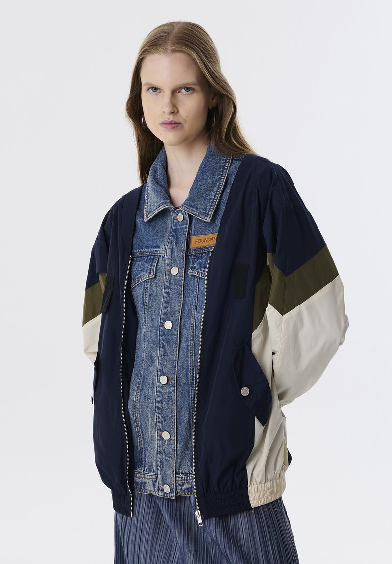 Marineblauwe jas met denim voorkant, olijf- en crèmekleurige accenten. Voorzien van drukknoopsluiting, grote kraag en ritszakken. Lichtgewicht stof, ontspannen pasvorm.