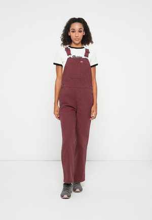 Salopette bordeaux con spalline regolabili, tasca frontale e design loose-fit, abbinata a una t-shirt bianca con bordi neri e scarpe grigie.