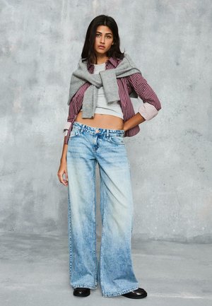 Junge Frau in weit geschnittenen, ausgewaschenen Jeans, grauem, bauchfreiem Oberteil, bordeauxrot gestreiftem Hemd mit hochgekrempelten Ärmeln, grauem Pullover über den Schultern und schwarzen Schuhen.