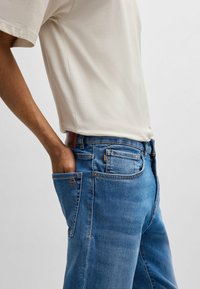 Béžová košeľa s krátkym rukávom v kombinácii s modrými rifľovými džínami. Džínsy majú oranžové a strieborné akcenty, predné zapínanie na gombík a podrobné prešívanie.