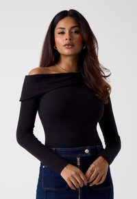 Zwarte off-shoulder top met lange mouwen, een aangesloten pasvorm en een gedrapeerde halslijn. Model draagt een gouden ketting en een blauwe spijkerrok.