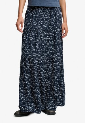 Personne portant une longue jupe bleu marine avec de petits motifs floraux blancs, des chaussures noires et un t-shirt bleu, debout devant un fond uni.