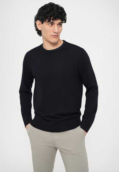 Granatowy sweter z dzianiny z okrągłym dekoltem, ściągaczami na rękawach i u dołu, noszony z jasnobeżowymi spodniami, z subtelnym zielonym akcentem przy dekolcie.