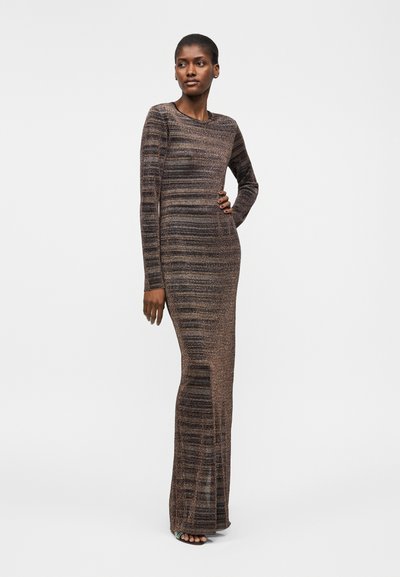 Γυναίκα στέκεται με το ένα χέρι στη μέση, φοράει μακρυμάνικο, μακρύ, ριγέ καφέ πλεκτό φόρεμα και ανοιχτά πέδιλα.