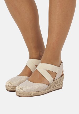 Alpargatas de cuña con suela de yute trenzado, parte superior de tela beige y tiras elásticas entrecruzadas. Punta redondeada con detalles cosidos.