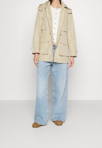 Beige parka-jacka med huva, två framfickor och silverdetaljer, i kombination med vida ljusblå jeans och bruna skor.