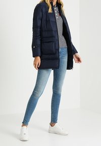 Námořnická prošívaná bunda s kapsami vpředu a zipem. Nošena přes pruhované tričko a doplněna světle modrými džíny a bílými teniskami.