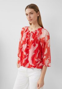 Blouse à motifs rouges et roses avec une coupe décontractée, des manches trois quarts et un col froncé. Fabriquée en tissu léger et transparent.