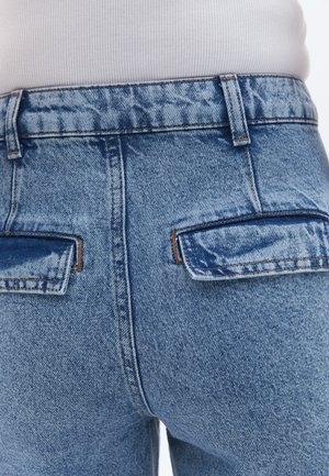 Světle modré džíny s vysokým pasem, které mají dvě zadní kapsy, jemné vyblednutí a kontrastní prošívání podél pásku a kapes.