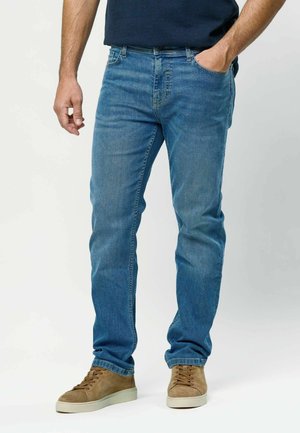 Bărbat îmbrăcat în blugi albaștri și adidași maro din piele întoarsă, cu o mână în buzunar și cealaltă relaxată pe lângă corp.