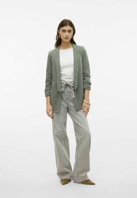 Lichtgroene blazer met een zachte textuur, witte getailleerde blouse en hooggetailleerde, wijde grijze broek. Bevat subtiele details met elastische manchetten.