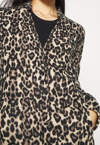 Kobieta w leopardzim płaszczu noszącym czarną bluzkę z delikatnym złotym łańcuszkiem, z ręką schowaną w kieszeni płaszcza.