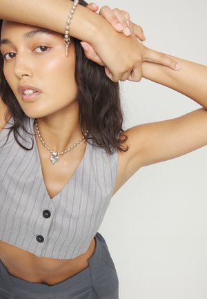 Joven con tono de piel medio, vestida con un chaleco gris a rayas y un collar plateado en forma de corazón, posando con los brazos levantados cerca de la cabeza.