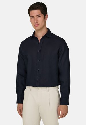 Homme portant une chemise noire à manches longues boutonnée, rentrée dans un pantalon beige taille haute, debout devant un fond uni.