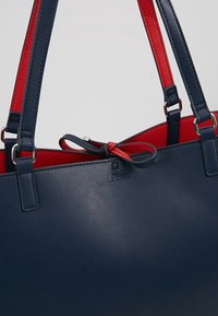 Niebieska torebka z syntetycznej skóry z czerwoną obiżką, dwoma mocnymi uchwytami, wytłoczonym logo z przodu i detalem kokardy przy zapięciu.