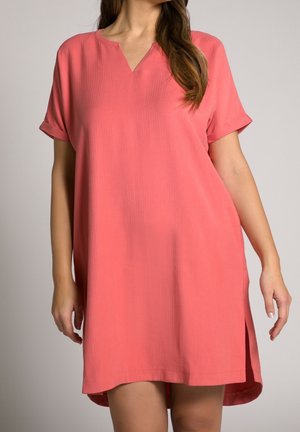Kvinde iført koralpink kjole med korte ærmer, V-hals og sideslids, stående mod en ensartet lys baggrund.
