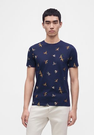 Jeune homme aux cheveux courts noirs portant un t-shirt bleu marine avec des imprimés colorés d'oiseaux et un pantalon beige clair sur un fond uni.