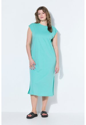 Kobieta stojąca, ubrana w turkusową sukienkę midi z krótkimi rękawami i czarne sandały typu klapki na jednolitym białym tle.