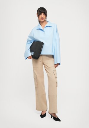 Woman wearing light blue oversized shirt, beige cargo pants, and black heels holding a black laptop sleeve against a white background.