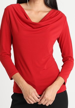 Femme portant un haut rouge ajusté à manches longues avec un décolleté drapé et un pantalon noir, mains jointes devant.