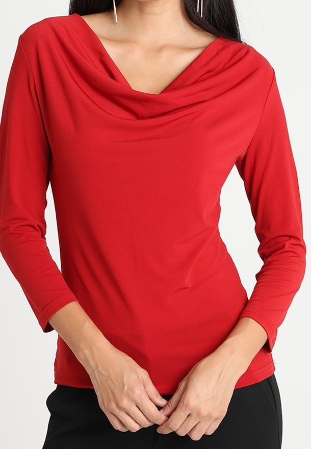 Femme portant un haut rouge ajusté à manches longues avec un décolleté drapé et un pantalon noir, mains jointes devant.