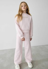 Rosa sweatshirt och matchande byxor i mjukt material, med guldfärgad text "BAKER." Avslappnad passform med ribbade ärmslut och vid byxdizajn.