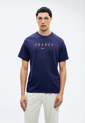 Άνδρας που φοράει μπλε σκούρο μπλουζάκι με την επιγραφή «FRANCE» και λογότυπο Nike σε μπλε χρώμα, συνδυασμένο με ανοιχτό γκρι παντελόνι, στέκεται μπροστά σε λευκό φόντο.