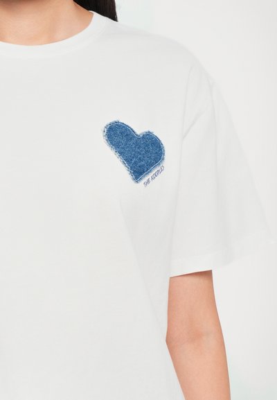 Valge puuvillane t-särk, millel on sinine denim süda vasakul rinnal, narmendavate servadega ja allpool südametätoveeringut on tekst "THE KOOPLES".