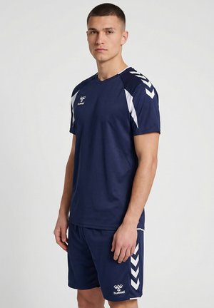 Junger Mann, der navyblaue hummel-Sportkleidung mit weißen Chevron- und Logodetails trägt und vor einem schlichten hellen Hintergrund steht.