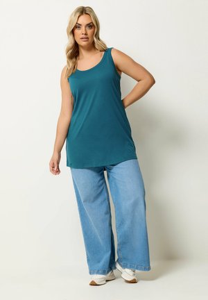 Blonde vrouw die een teal mouwloze top met brede schouderbanden draagt, lichtblauwe wijde broekspijpen en witte sneakers, staand tegen een lichte achtergrond.