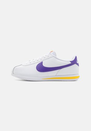 Baskets blanches basses avec un swoosh Nike violet et un onglet de talon, une bande jaune sur la semelle et des lacets blancs.
