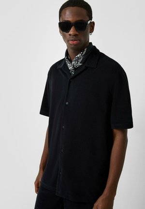 Homme portant une chemise noire à manches courtes boutonnée, des lunettes de soleil noires, des boucles d'oreilles créoles et une écharpe à motifs noirs et blancs sur un fond gris uni.