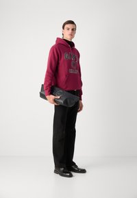 Felpa bordeaux con logo, jeans neri e scarpe nere. Il modello tiene in mano una borsa nera con texture e accenti arrotondati. Sfondo semplice.