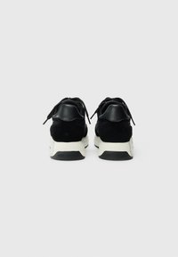 Baskets noires en daim et cuir avec une semelle blanche, dotées de lacets et d'un talon marqué. Design minimaliste avec des matériaux contrastés.