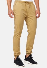 Pantalon de jogging beige avec ceinture élastique et cordon de serrage. Bas de jambes effilés. Porté avec des chaussures de sport noires. Texture lisse, sans motifs.