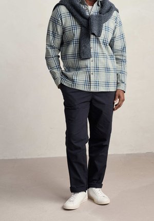 Hombre con camisa de cuadros azul y blanca, suéter gris sobre los hombros, pantalones oscuros y zapatillas blancas, de pie en el interior.