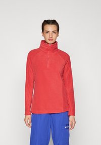 Pullover in pile rosso con colletto alto e design a zip quart. Presenta maniche lunghe e una texture morbida. Abbinato a pantaloni cargo blu.