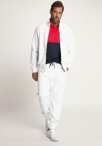 Chaqueta blanca con cremallera, camiseta polo a rayas en rojo y azul marino, pantalones de chándal blancos y zapatillas ligeras. El conjunto presenta un diseño deportivo y casual.