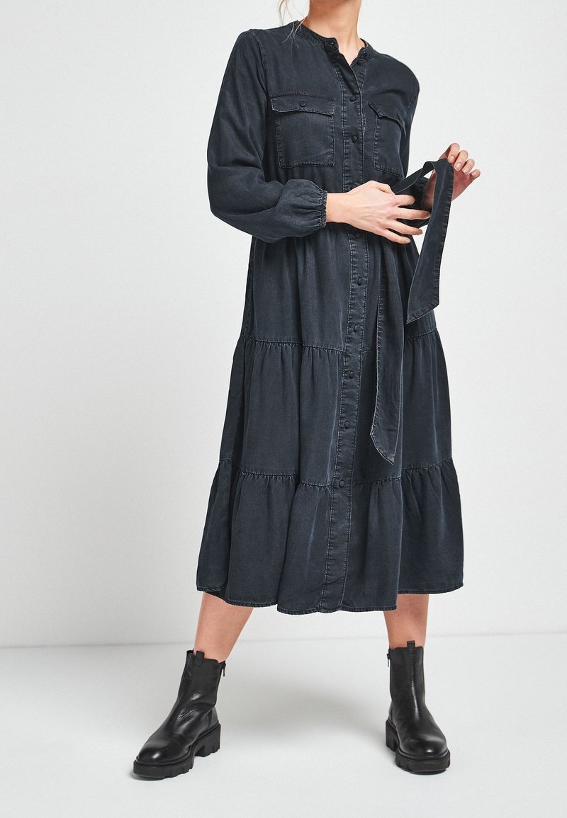 Femme portant une robe midi noire à manches longues boutonnée, tenant une ceinture en tissu assorti, associée à des bottines noires sur un fond uni.