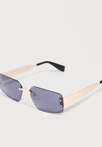 Λεπτά χρυσά μεταλλικά γυαλιά ηλίου με ορθογώνιους σκούρους φακούς και μαύρες καμπύλες άκρες βραχίονα, ξεκουρασμένα σε μια ανοιχτόχρωμη επιφάνεια.