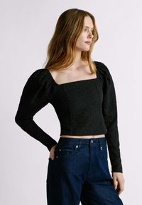 Top corto nero con texture, scollatura quadrata e maniche lunghe a sbuffo, abbinato a pantaloni in denim blu scuro. Nota il tessuto liscio e la vestibilità sagomata.