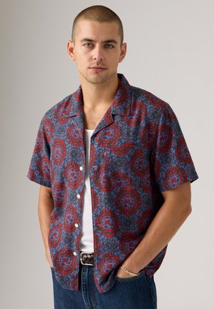 Junger Mann mit kurzen Haaren, der ein rot und blau gemustertes kurzärmliges Hemd, ein weißes Tanktop, eine blaue Jeans und einen geflochtenen Gürtel trägt, mit den Händen in den Taschen.